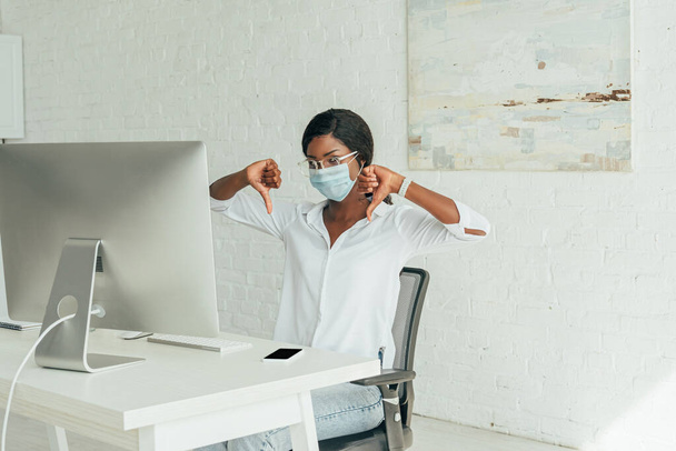 disgustado afroamericano freelancer mostrando los pulgares hacia abajo mientras está sentado cerca del monitor de la computadora en casa
 - Foto, Imagen