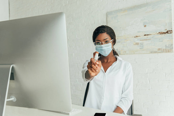 young african american freelancer in medical mask showing antiseptic spray while sitting near computer monitor at home - Φωτογραφία, εικόνα