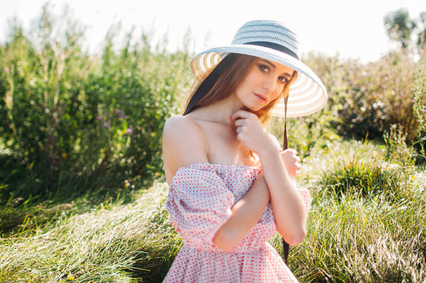a beautiful girl in a pink dress and a hat with a brim at sunset in a blooming green field. look at the camera. romantic spring image - Foto, imagen