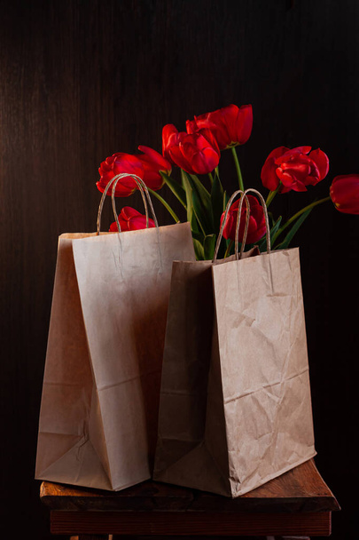 Kraft paper bags with blooming red tulips bouquet on black background. Dark moody low key minimalism style flowers and food delivery packaging banner copy space mockup. Donations help recycling boxes. - Zdjęcie, obraz