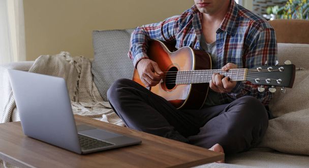 Music college hipster student in checkered plaid shirt practicing acoustic guitar exercise, reading notes from laptop computer. Man taking an online musical courses at home during quarantine. Background, close up - Photo, image