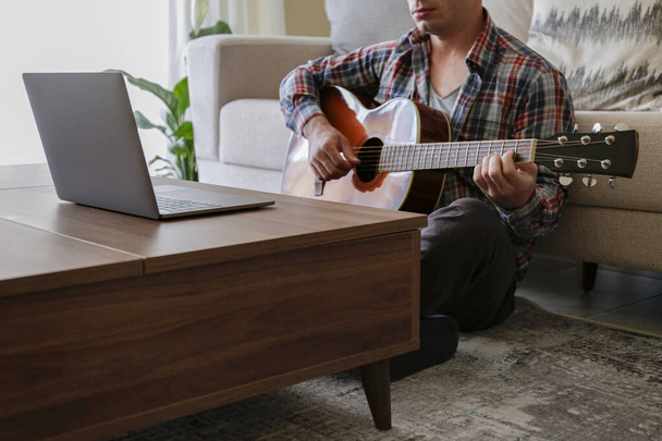 Music college hipster student in checkered plaid shirt practicing acoustic guitar exercise, reading notes from laptop computer. Man taking an online musical courses at home during quarantine. Background, close up - Фото, изображение