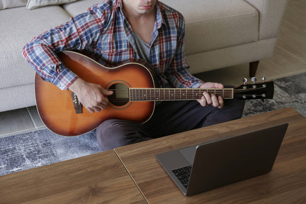 Muziek college hipster student in geruite geruite shirt oefenen akoestische gitaar oefening, het lezen van notities van laptop computer. Man die thuis een online muziekcursus volgt tijdens quarantaine. Achtergrond, close-up - Foto, afbeelding