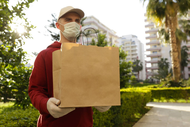 Delivery guy wearing red hoodie, disposable gloves and mask for coronavirus protection, holding blank brown paper grocery bag outside of high rise apartment building. Close up, copy space. - Photo, Image