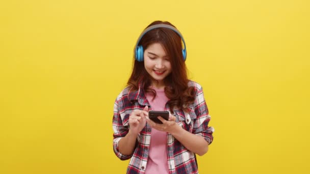 Молодая азиатка в беспроводных наушниках слушает музыку со смартфона с веселым выражением лица в повседневной одежде и смотрит на камеру на желтом фоне. Концепция выражения лица. - Кадры, видео