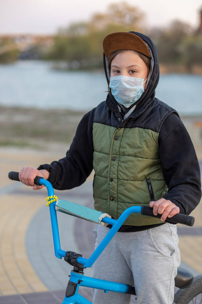 Porträt eines Teenagers im Sonnenuntergang mit Maske auf einem blauen Fahrrad. Quarantäne. Es ist verboten, mit dem Fahrrad zu fahren. Covid-19. - Foto, Bild
