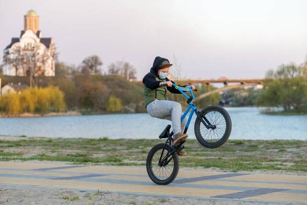 Zamaskowany nastolatek na niebiesko-czarnym rowerze jeździ na tylnym kole. Nabrzeże rzeki. Kościół i most w tle. Kowid-19. - Zdjęcie, obraz