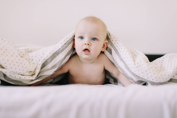 Beyaz battaniyenin altında saklanan bebek portresi. 8 aylık tatlı bebek yatakta emekliyor. Mide zamanı. Çocuklar için tekstil ve yatak - Fotoğraf, Görsel