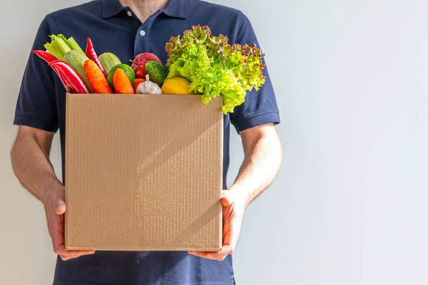 Food delivery service - man with a cardboard box of fresh vegetables on a gray background. Copy space - Fotoğraf, Görsel