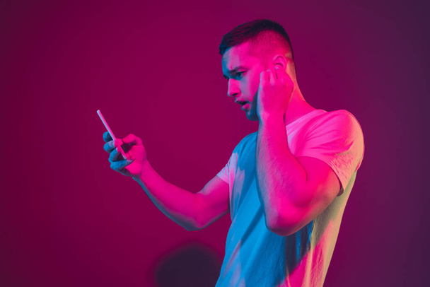 Caucasian mans portrait isolated on pink-purple studio background in neon light, with smartphone and earphones - Foto, Imagem