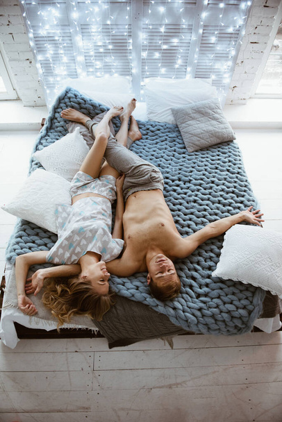 Couple in the bedroom on the bed in the bedroom. A blonde girl in pajamas, a guy with a naked torso. Love and hugs. A beautiful couple. New Year's interior. Christmas time. A blanket of large mating. - Foto, Imagem