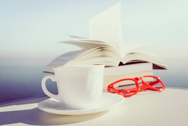 Reading on holidays. Coffee cup and books on table outdoors against blue sea water background. Book pages fluttering in the wind. - Photo, Image