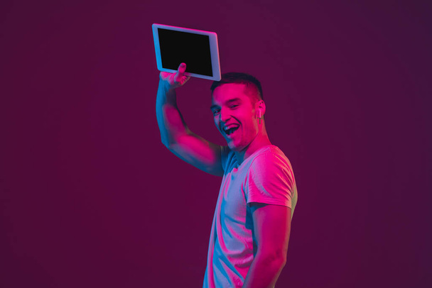 Caucasian mans portrait isolated on pink-purple studio background in neon light, using tablet - Фото, изображение