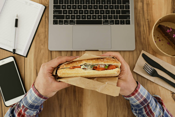 Lunch break concept. Man eating takeout food by his workspace with laptop, blank screen phone & notebook. Panini sandwich, chocolate cake. Top view, POV, close up, copy space, wooden table background. - Foto, Imagen