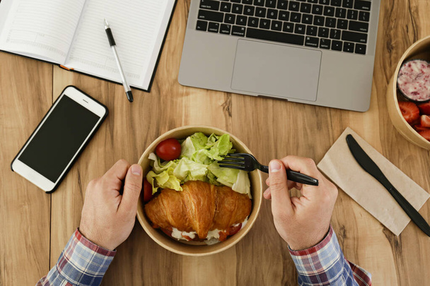 Lunch break concept. POV shot of man eating takeout food by his workspace. Laptop, blank screen phone & notebook. French croissant with salad. Top view, close up, copy space, wooden table background - Photo, image