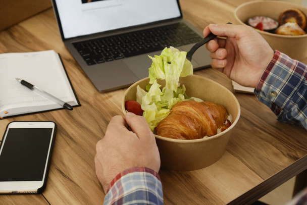 Lunch break concept. POV shot of man eating takeout food by his workspace. Laptop, blank screen phone & notebook. French croissant with salad. Top view, close up, copy space, wooden table background - Photo, image