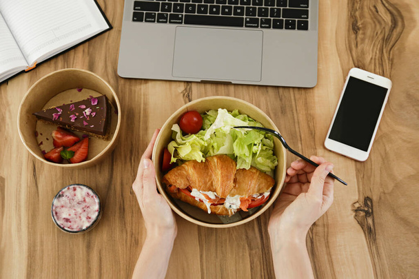 Lunch break concept. Woman eating takeout food by her workspace. Laptop, blank screen phone & notebook. French croissant, salad, chocolate cake. Top view, POV, close up, copy space, wooden background - Foto, Imagen