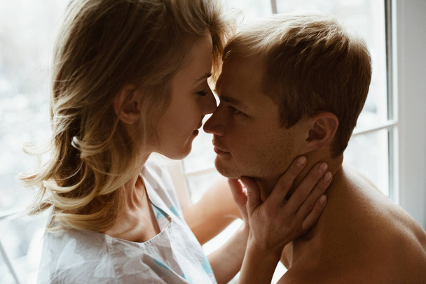Stylish interior. A couple near the window, under one blue blanket of large mating. Gentle embrace and kisses. Blonde. Floor lamp. Athletic build. Gray pajamas in hearts. Love story. - Fotografie, Obrázek