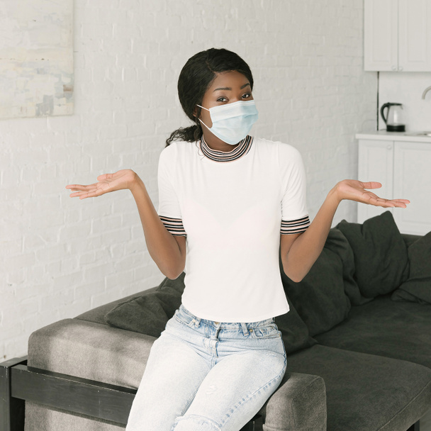 confused african american woman in medical mask showing shrug gesture while looking at camera - Фото, изображение