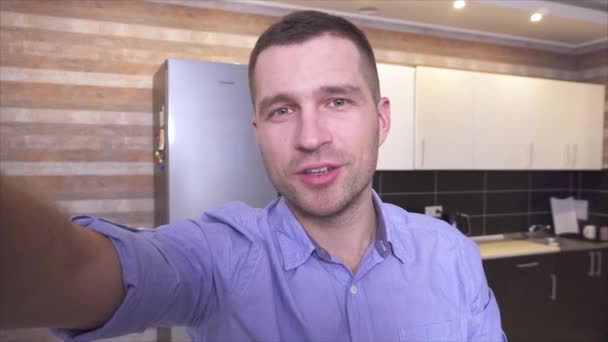Young male blogger recording video at home. Hold camera with hand and talk. Point on back kitchen furniture. Talk cheerful and confident. - Footage, Video