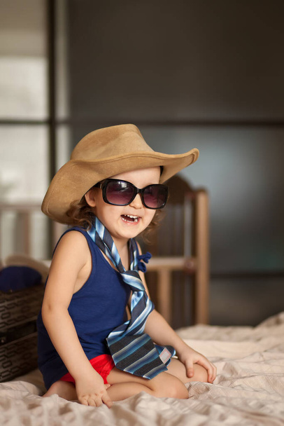 Happy childhood. Cute girl playing in a fashion, wearing big cowboy hat and sunglasses. Adorable child having fun indoors. Quarantine fun. - Фото, изображение