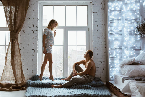 Glowing New Year garlands. Stylish interior. couple near window, under blue blanket of large mating. Gentle embrace and kisses. Blonde. Floor lamp. Athletic build. Gray pajamas in hearts. Love story. - Foto, Imagem