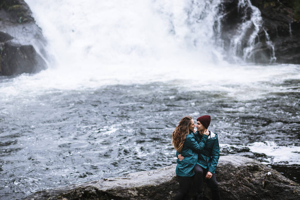 A couple of lovers in green raincoats, sitting on a rock, against the backdrop of a waterfall - Photo, image
