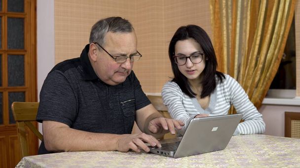 Hija enseña a padre cómo utilizar un ordenador portátil. Una joven le muestra a su padre dónde hacer clic en un ordenador. La familia está sentada en una habitación cómoda
. - Foto, imagen