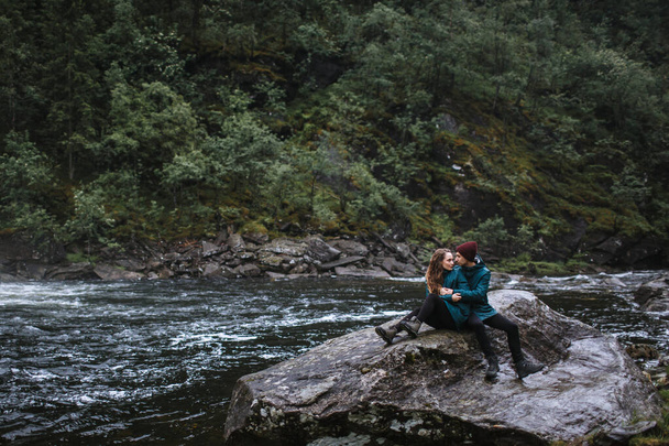 A couple of lovers in green raincoats, sitting on a rock, against the backdrop of a waterfall - Foto, Bild
