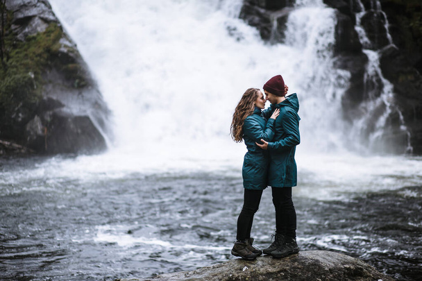 Portrait of lovers in green raincoats, standing on a stone, against the background of a waterfall - Foto, immagini