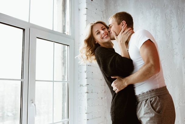 A pair of lovers, hug, kiss, laugh and have fun. The girl in a long black sweater and white high golf. A guy in a white T-shirt and trousers. Light gray interior, large window, brown stylish chair. - Фото, изображение