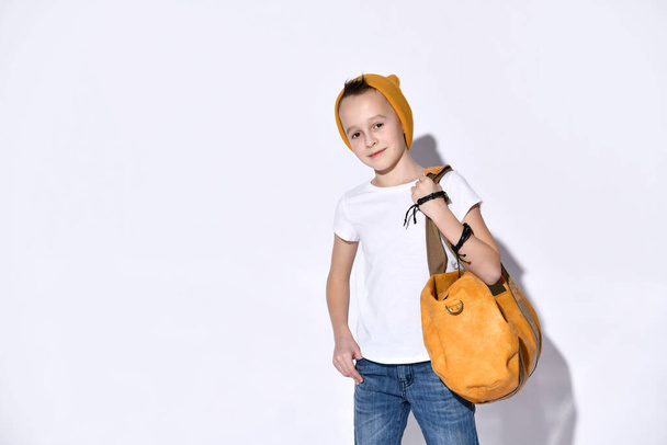 Portrait A teenage child with an orange travel bag, wearing a hat, sunglasses, a T-shirt, a blue denim suit, black sneakers and a bracelet. - Fotografie, Obrázek