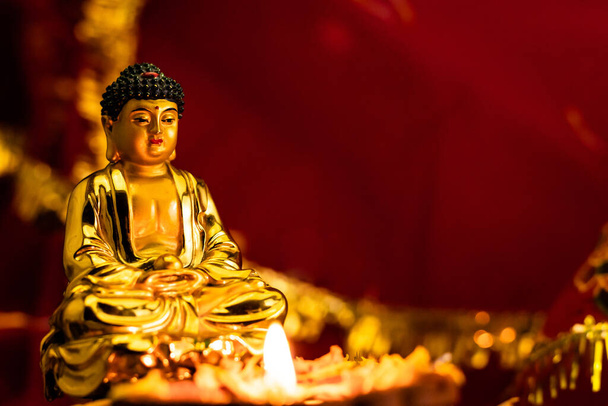 közeli kis buddha szobor izzó diya ellen elmosódott piros arany háttér. buddhizmus és hit fogalma - Fotó, kép