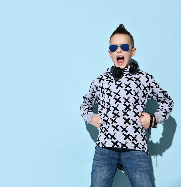 Teenage kid with headphones on neck, in sunglasses, blue jeans and stylish hoodie. He calling someone, posing on blue background - Foto, afbeelding