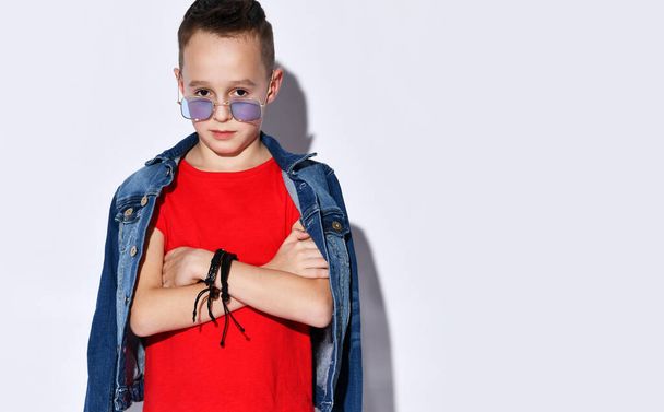 Teenage kid in purple sunglasses, red t-shirt and denim jacket, black bracelet. Hands folded, posing isolated on white background - Foto, immagini