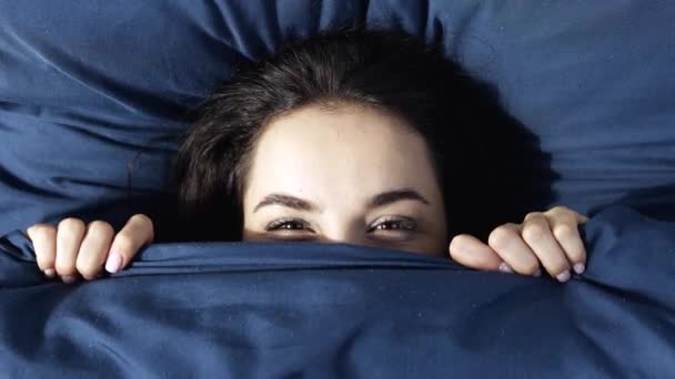 Jonge vrouw blijft thuis tijdens quarantaine. Vrolijk positief meisje liggend onder donkerblauw linnen deken. Verschijn en kijk op camera met een glimlach. Verborgen gezicht onder deken. - Video