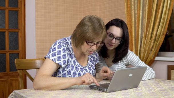 La hija le enseña a la madre a usar una computadora portátil. Una joven le muestra a su madre dónde hacer clic en un ordenador. La familia está sentada en una habitación cómoda. Mujer mirando a la cámara y mostrando el pulgar
 - Foto, Imagen