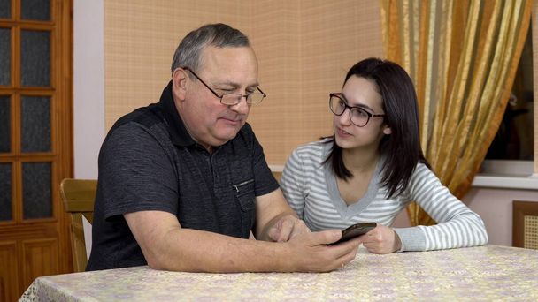 Дочка вчить батька користуватися телефоном. Молода жінка показує своєму старому батькові, де можна натиснути на смартфон. Чоловік зателефонував по телефону. Сім'я сидить у зручній кімнаті
. - Фото, зображення