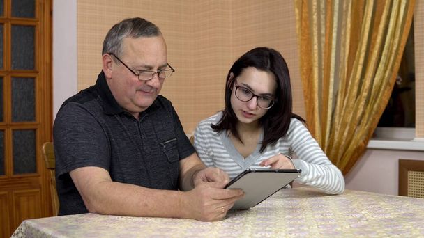Una hija le enseña a su padre a usar una tableta. Una joven le muestra a su viejo padre dónde hacer clic en una tableta. La familia está sentada en una habitación cómoda
. - Foto, imagen