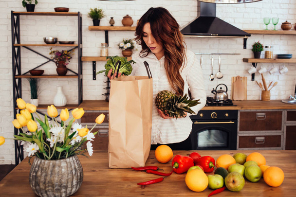 A young woman came back from a supermarket. There are many fruits and vegetables: apples, red peppers, hot peppers, oranges, pears in her bag. She is keeping pineapple and broccoli in hands. Kitchen. - Foto, afbeelding