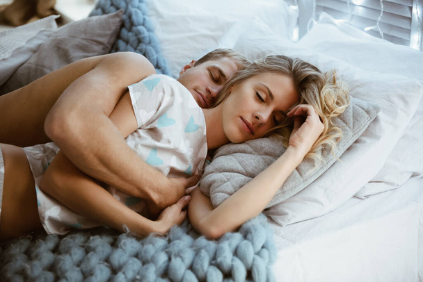 Couple in the bedroom on the bed in the bedroom. A blonde girl in pajamas, a guy with a naked torso. Love and hugs. A beautiful couple. New Year's interior. Christmas time. A blanket of large mating. - Photo, Image