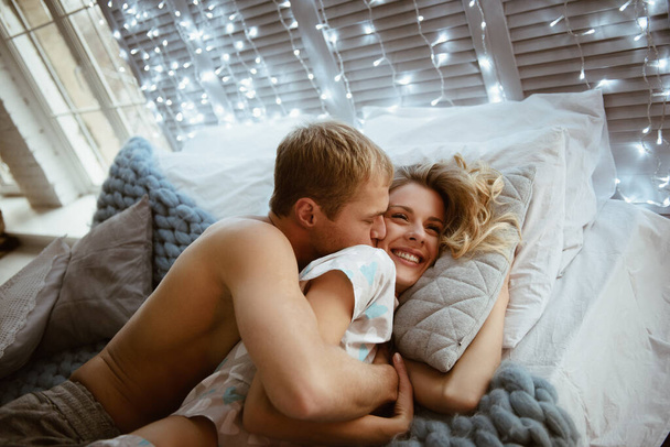 Couple in the bedroom on the bed in the bedroom. A blonde girl in pajamas, a guy with a naked torso. Love and hugs. A beautiful couple. New Year's interior. Christmas time. A blanket of large mating. - Photo, Image