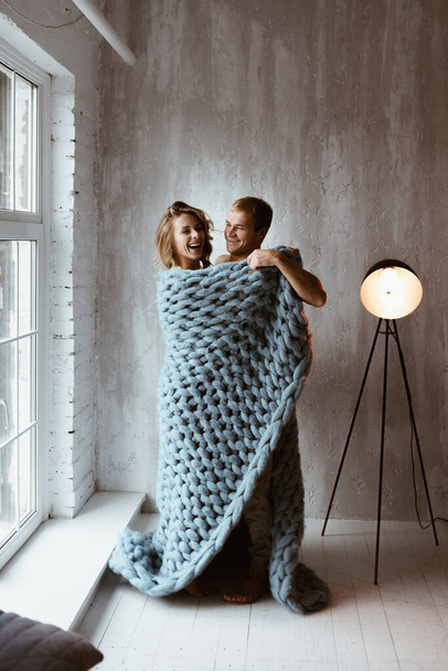 Stylish interior. A couple near the window, under one blue blanket of large mating. Gentle embrace and kisses. Blonde. Floor lamp. Athletic build. Gray pajamas in hearts. Love story. - Photo, Image