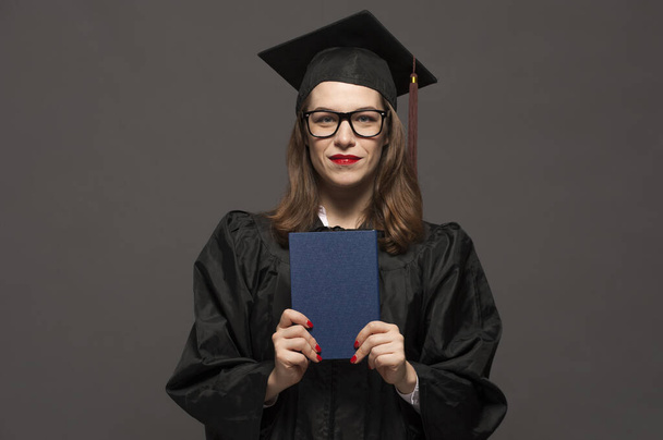Charming female student wearing black mantle and standing with diploma in hands - Фото, изображение