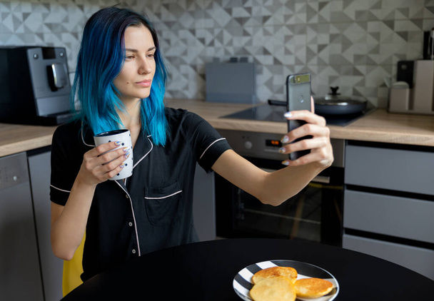Beautiful young hipster woman taking selfie at modern kitchen. Social media communication quarantine concept - 写真・画像