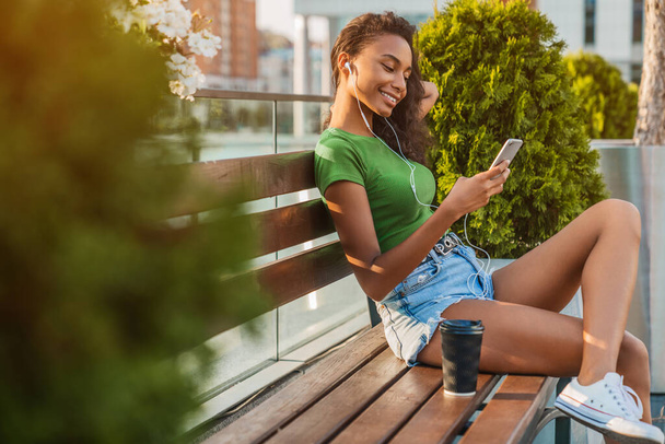 Pretty young woman using smartphone with earphones while sitting on street bench - Foto, Imagem
