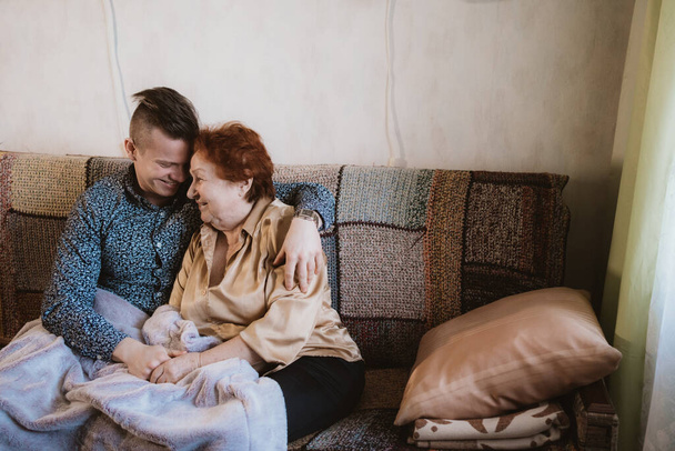 kleinzoon zitten met oma op de bank onder de deken en knuffelen haar, familie liefde - Foto, afbeelding