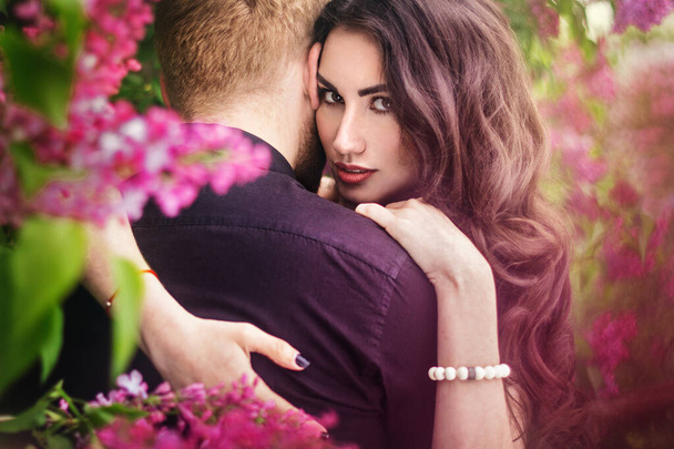 Menyasszony és vőlegény közel orgonafához. Egy szerelmes pár ölelkezik egy virágzó tavaszi kertben. Egy nő lila ujjatlan ruhában, finom nyaklánccal a nyakában. Vörös hajú férfi fekete öltönyben megcsókol és megölel egy lányt. Esküvő a gyönyörű parkban. - Fotó, kép
