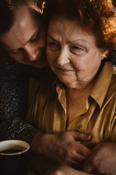 kleinzoon die zijn grootmoeder knuffelt, aan tafel zit, kleinzoon die grootmoeder bezoekt, steun van de oudere generatie, foto met warm liht - Foto, afbeelding