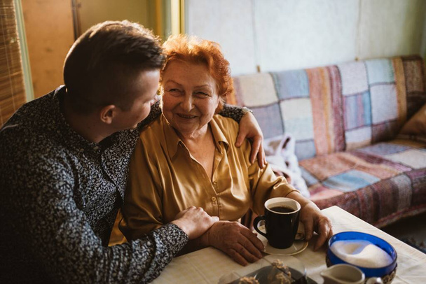 Enkel umarmt Großmutter, sitzt am Tisch, Enkel besucht Großmutter, Unterstützung der älteren Generation, Foto mit warmem Licht - Foto, Bild